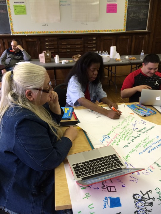 Diane seated at a transition planning event, working with two young people. There is scroll paper and markers on the table. George is in the background.