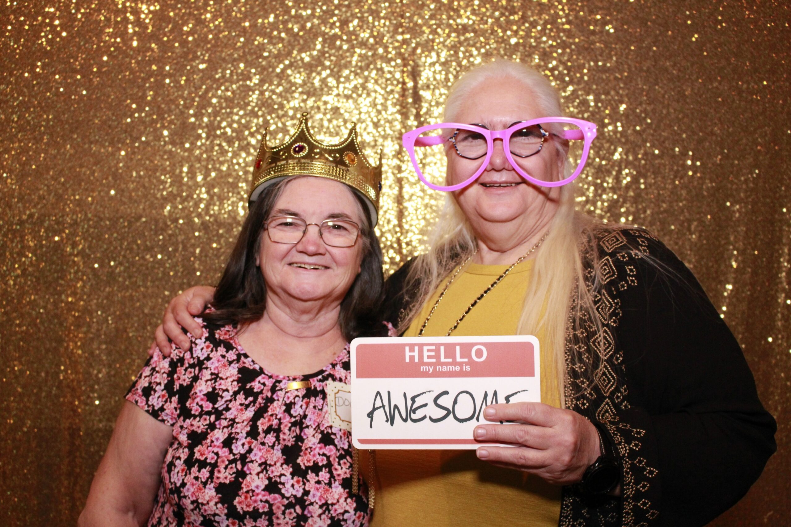 Diane and Doris posing. Doris is wearing a tiara, and Diane is wearing silly glasses holding a sign that says “Hello my name is Awesome”