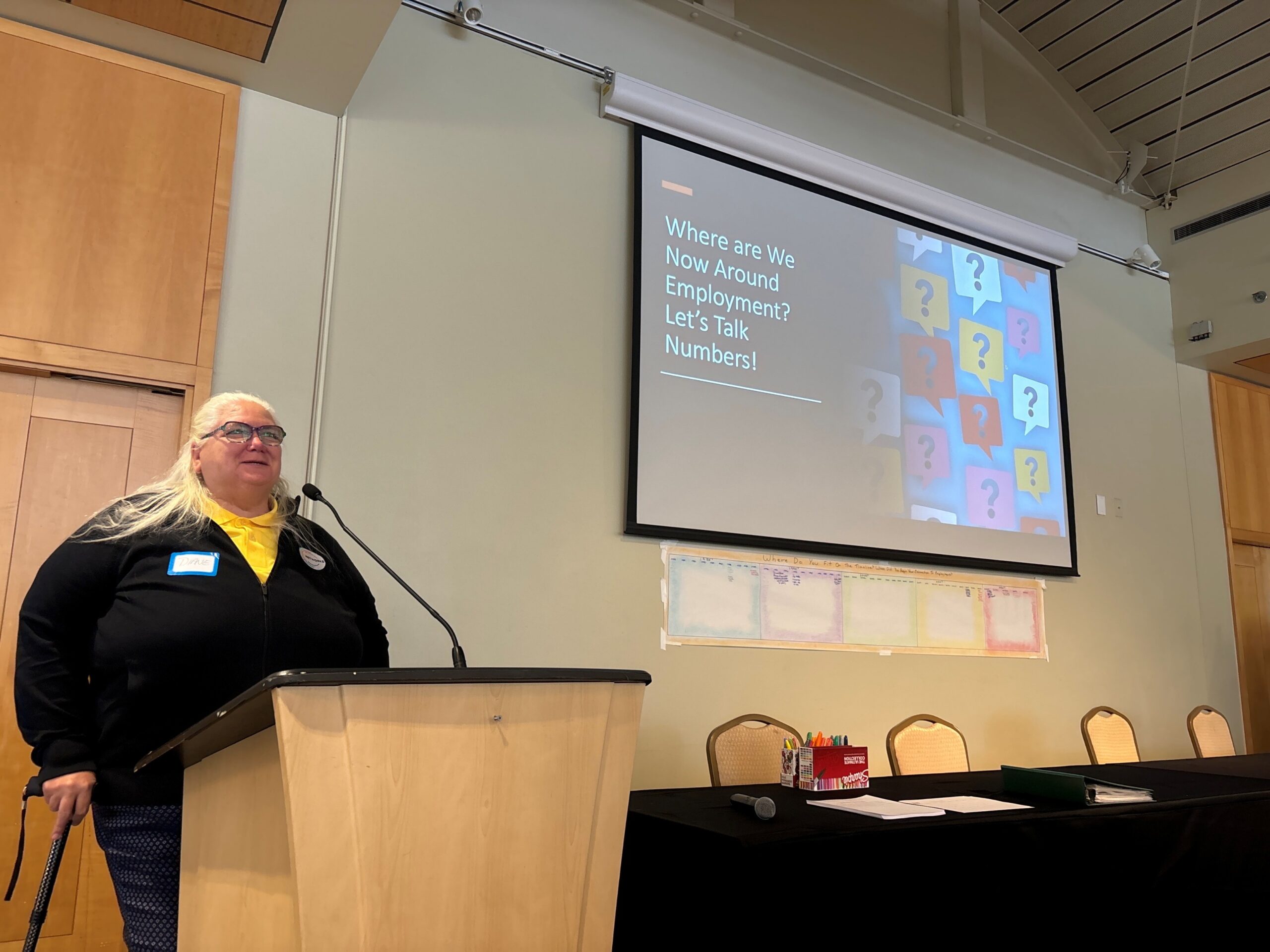 Diane is presenting at an employment stakeholder event at the Independent Visitors Center. The slide in the background reads “Where are we now around employment? Let’s talk numbers”